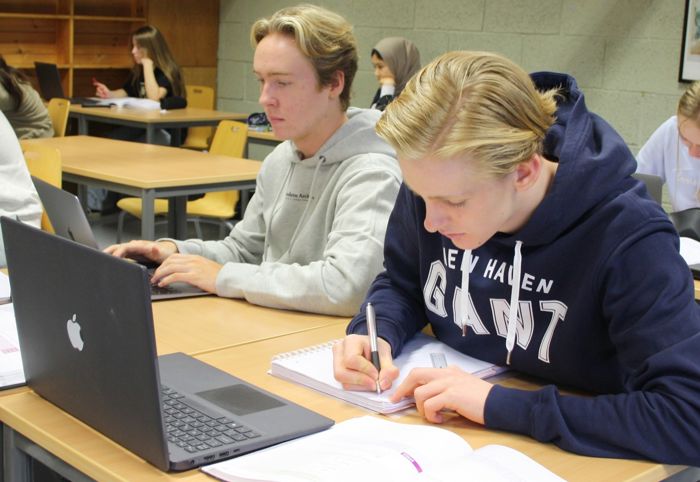 Bilde av to gutar som sit ved ein pult i eit klasserom, den eine jobbar på PC og den andre skriv i ei notatbok.